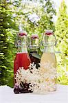 Three glass bottles of homemade syrup on a garden table: rhubarb syrup, cherry syrup and elderflower syrup