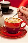 A hand stirring a hot cappuccino decorated with a heart