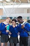 Three happy students wearing school uniforms using smartphone in school corridor