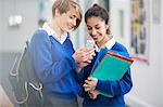 Two smiling female students wearing school uniforms looking at smartphone in school corridor