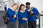 Three smiling students wearing blue school uniforms with mobile phone in school corridor
