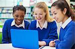 Three smiling female students wearing blue school uniforms working on laptop in library