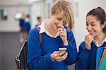 Two smiling female students looking at mobile phone in school corridor