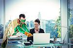 Superhero helping businesswoman working at office desk