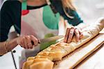 Close up of a woman wearing an apron, sitting at a table, slicing a baguette.