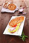 Wholegrain baguette with ham and fresh vegetables, decorated with onion and rosemary on brown wooden background.