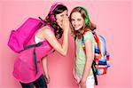 Trendy schoolgirls sharing a secret with each other. All on pink background