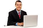 Male executive at work desk operating laptop and smiling camera
