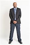 Full length friendly Indian businessman in formal suit looking at camera, standing on plain background.