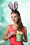 Pretty, happy blonde woman with long hair and rabbit ears on the head. She is holding bucket of little Easter eggs.