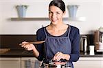 Young woman cooking the food for dinner over the stove in her kitchen standing holding the lid of a stainless steel saucepan and wooden ladle as she smiles at the camera