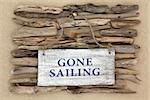 Gone sailing old weathered sign on driftwood and beach sand background.