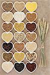 Grain and cereal food selection in heart shaped porcelain bowls over rough brown paper background with wheat ears.