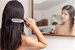 Young woman standing looking at her reflection in the bathroom mirror while brushing her wet hair with a hairbrush after washing it, view from behind