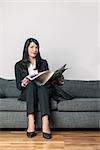 Attractive businesswoman in a stylish slack suit sitting reading a business journal or newspaper on a grey sofa with her briefcase alongside her as she waits for an appointment