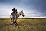 A cowboy standing leaning on a fence post on the range. A grey horse behind him.