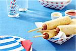 Homemade corn dogs in a paper serving container with paper plates, napkins, glasses and straws, studio shot on blue background