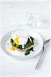 Poached egg with soft yolk and spinach on an english muffin, on plate with fork and glass of water, studio shot on white background