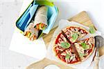 Homemade pizza on cutting board and healthy, vegetable sandwich wraps in container, studio shot on white background