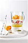 Protein fruit shake ingredients in a blender with empty glass and straw on a white cutting board, studio shot on white background