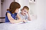 Girl reading book with brother at kitchen table