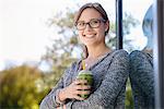 Portrait of young woman with takeaway coffee leaning against park building