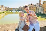 Happy mother and baby girl checking photos in camera on bridge overlooking ponte vecchio in florence, italy