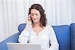 Pretty brunette speaking on the phone while using her laptop in the living room