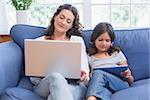 Happy mother and daughter sitting on the couch while using laptop and tablet in the living room