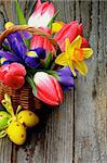 Arrangement of Yellow Daffodils, Magenta Tulips, Purple Irises in Wicker Basket with Yellow Easter Eggs closeup on Rustic Wooden background