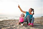 Healthy mother and baby girl sitting on beach in the evening and pointing
