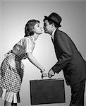 1950s 1960s HOMEMAKER WIFE IN CHECKED APRON KISSING BUSINESSMAN HUSBAND IN SUIT HAT AND TIE AS SHE HANDS HIM A BRIEFCASE