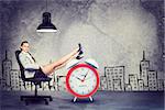 Businesswoman sitting in office chair with her feet up on alarm-clock, in front of raw concrete wall with sketch of city on it, looking at camera