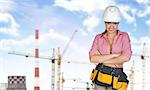 Woman in tool belt and helmet, crossed arms. Tower cranes, chimneys as backdrop