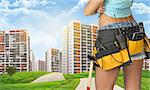 Woman in tool belt with different tools stands back. Cropped image. Green hills with road and buildings on background