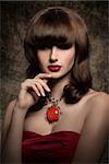 Pretty, mysterious, luxury, brunette woman with nice hairstyle with fringe covering her eyes, wearing red, satin top and big, old fashioned necklace, her lips are glossy and red.