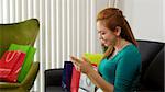 Young latina woman back at home after shopping, surrounded by bags on sofa. She holds a smartphone and types a post on social network