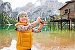 A young, blonde girl standing on the shores of Lake Bries is smiling while holding a digital camera up. In the background, the water provides a mirror reflection of the autumn scenery, including autumn leaves, the Dolomites, and a rustic wooden pier with a small house on the pier.