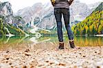 Study of the bottom half. A woman hiker stands at the edge of Lake Bries. In the foreground, a pebbly beach. In the background, the lake is a mirror reflection of the mountains and autumn colours.