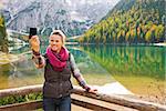 A smiling brunette taking a selfie on Lake Bries while leaning on a wooden guardrail. The still water reflects the autumn colours and the Dolomite mountains.