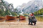 A smiling brunette mother kneels next to her daughter, pointing up. In the background, wooden boats float on the water, autumn colours, the Dolomite mountains, and forest.