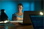 Beautiful woman working as interior designer, staying late at night in office with drawings and laptop computer to complete a project. The girl types a message on mobile phone.