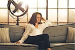 An elegant brunette is sitting on a sofa in a loft, pensive. In the background, a large window letting in light and an industrial standing fan.