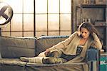 An elegant brunette woman wearing comfortable, casual clothing, leggings, and a cardigan is relaxing on a sofa in a loft. Sunlight shines through the window. Cozy atmosphere, industrial chic.