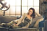 A brunette woman is smiling, relaxing on a sofa, feet curled under her, wearing comfortable clothing, leggings, and a cardigan. Industrial chic ambiance and cozy atmosphere.