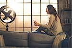 Seen from the side,a brunette woman is smiling, looking down at her phone sitting on the back of a sofa. Industrial chic ambiance and cozy atmosphere, sunlight is streaming through the loft window.