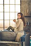 A brunette woman in comfortable clothing is is smiling and holding a hot cup of coffee, sitting on the back of a sofa. Industrial chic background, and cozy atmosphere. Loft decoration details.