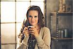 Head and shoulder shot of a brunette woman in comfortable clothing is smiling gently over the top of her coffee cup. She is casually dressed and in a cozy loft atmosphere. Urban chic decoration.