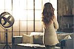 Seen from behind, a brunette woman in comfortable clothing is standing in a loft living room, looking out the loft window. Urban chic loft decoration details.