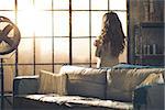 Looking away, a brunette woman in comfortable clothing is standing in a loft living room, hugging herself, looking out the window. Urban chic loft decoration details and window.